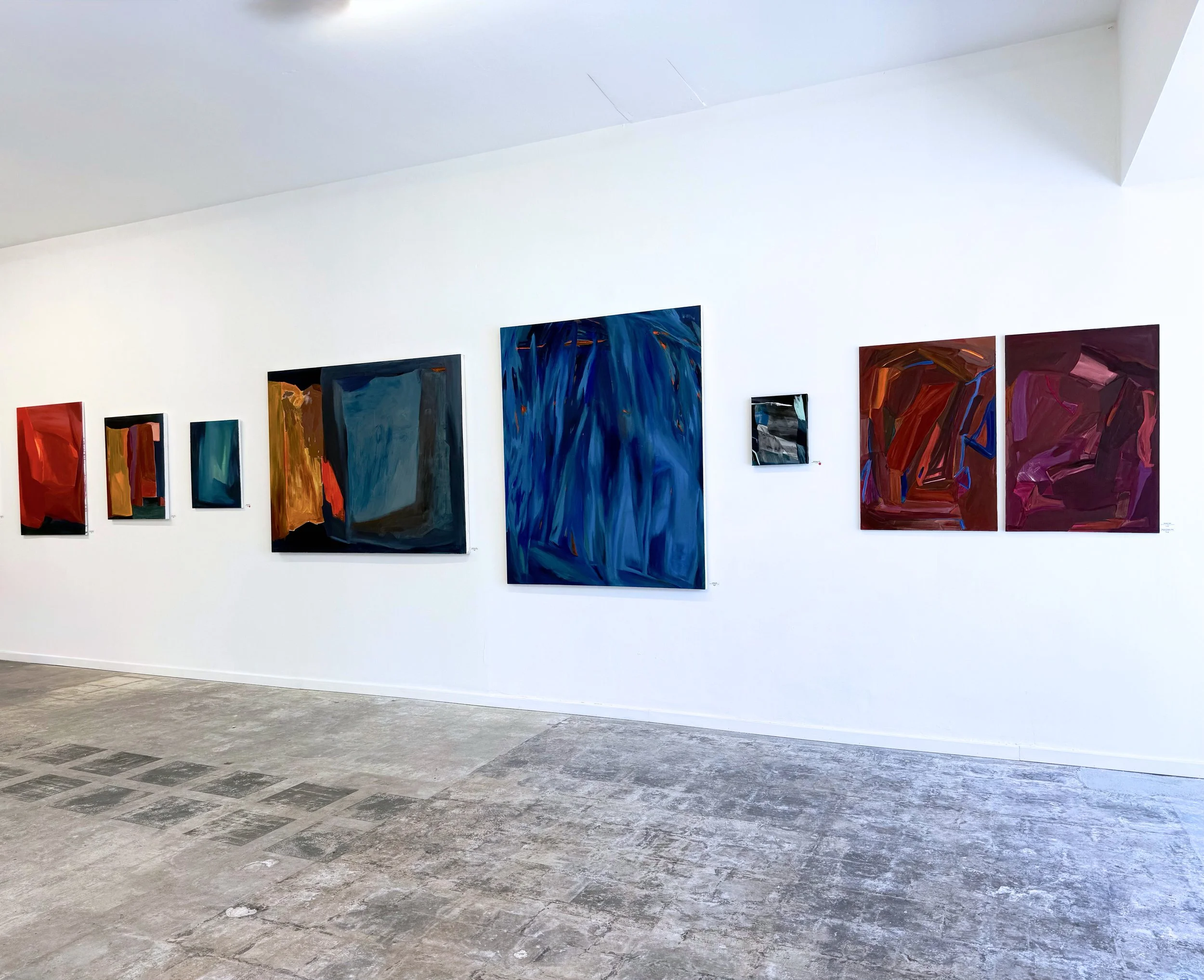 Gallery wall with abstract paintings in various colors, including red, blue, orange, and purple, displayed in an art museum with concrete floor.