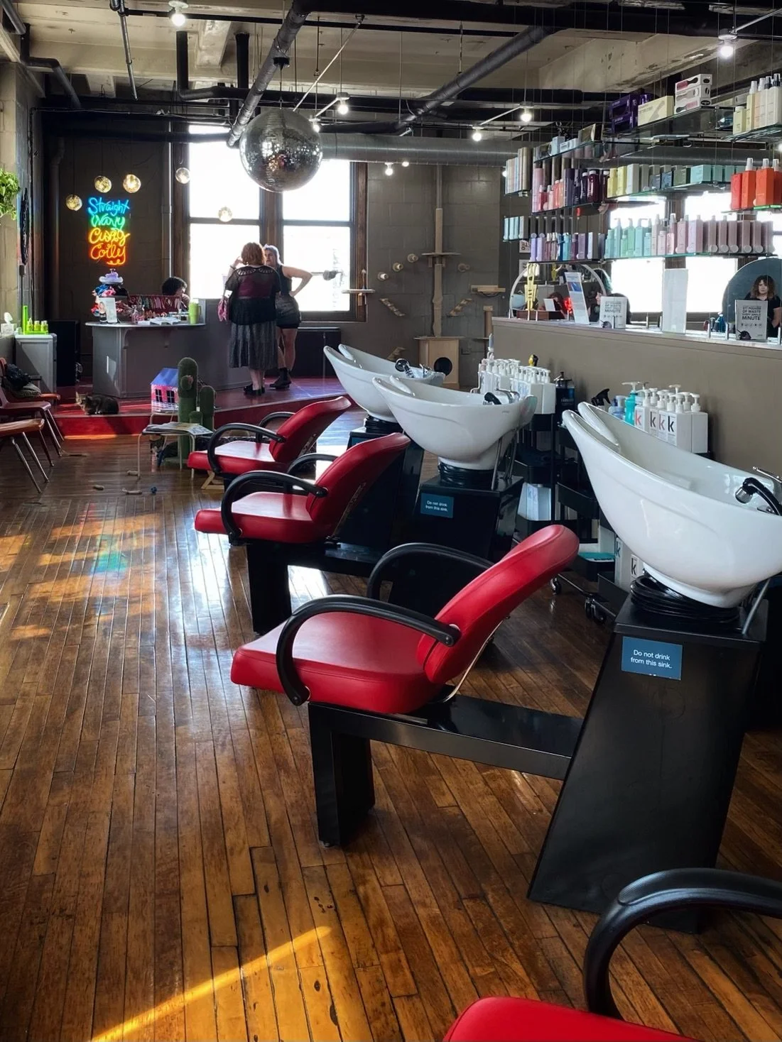 Salon with three white hair-washing sinks, red chairs, wooden floor, shelves with hair products, neon sign, and a small group of people near a window.