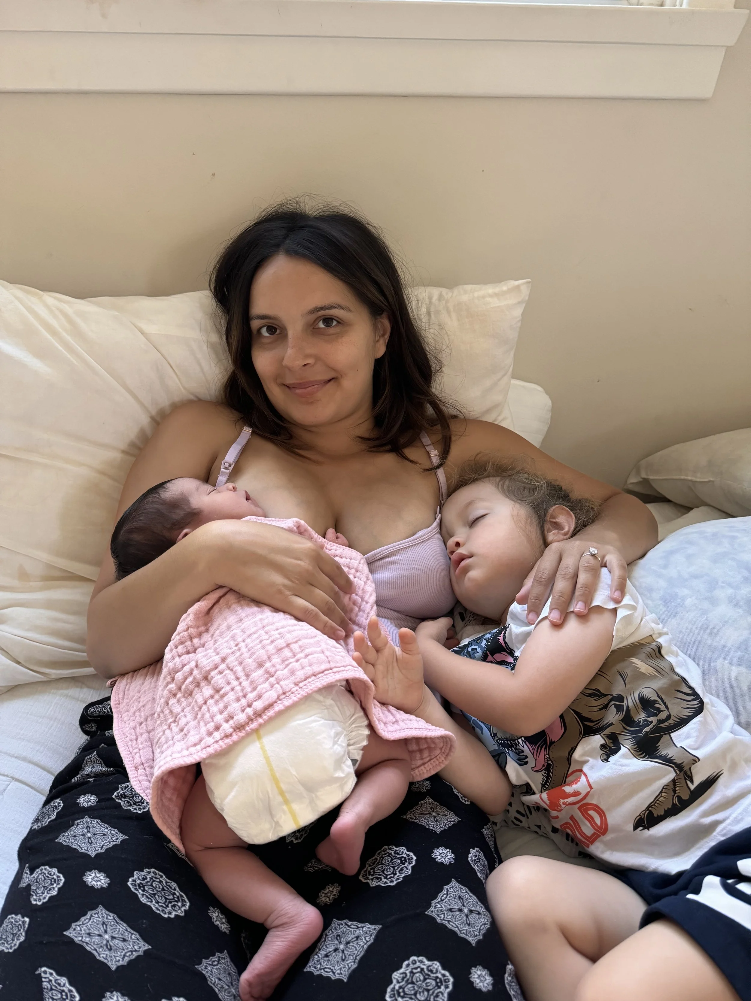 A woman lying on a bed, breastfeeding a newborn baby wrapped in a pink blanket, with a young child resting their head on her arm.