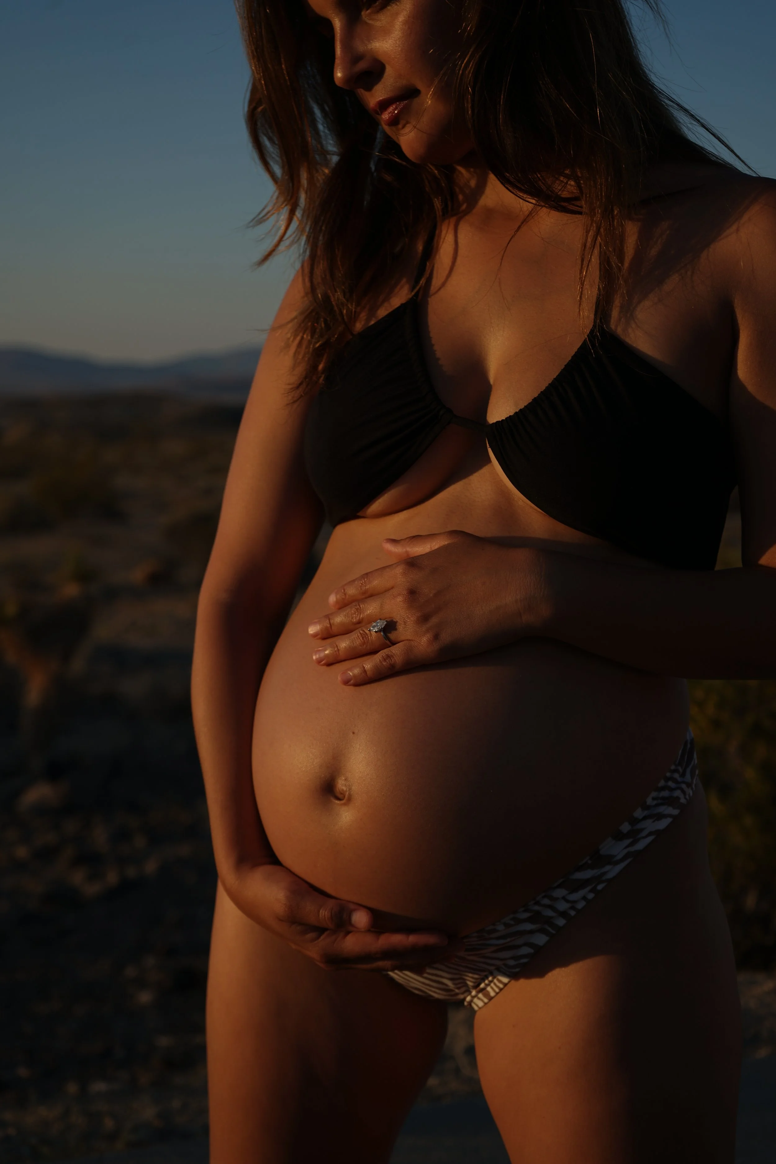 A pregnant woman in a black bikini top and striped bikini bottoms standing outdoors at sunset, holding her belly with both hands in a desert landscape.