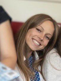 Young woman with light skin, light brown hair, earrings, and a nose ring smiling, sitting in what appears to be a restaurant or cafe with shelves and a large speaker in the background.