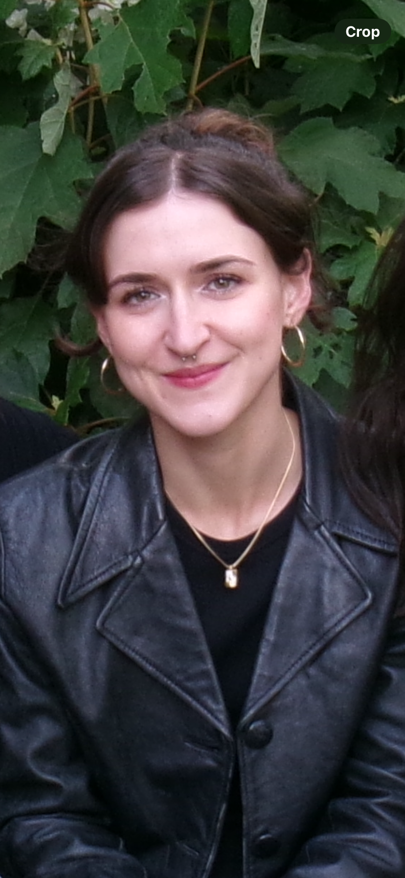 A young woman with shoulder-length wavy brown hair, wearing pearl earrings and a silver satin dress, smiling at the camera in front of a black metal fence.