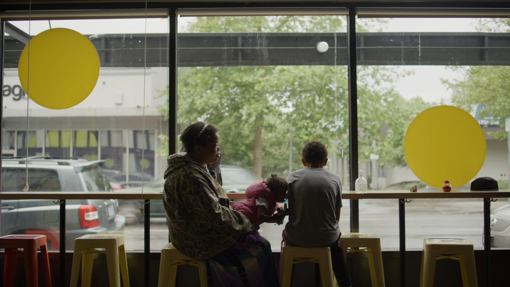 A woman and two children sit by a large window in a cafe, with the woman holding a young girl. The silhouettes of the people are visible against the outdoor view of parked cars and greenery. Yellow and red stools are in front of the window, decorated with large yellow circular ornaments hanging inside.