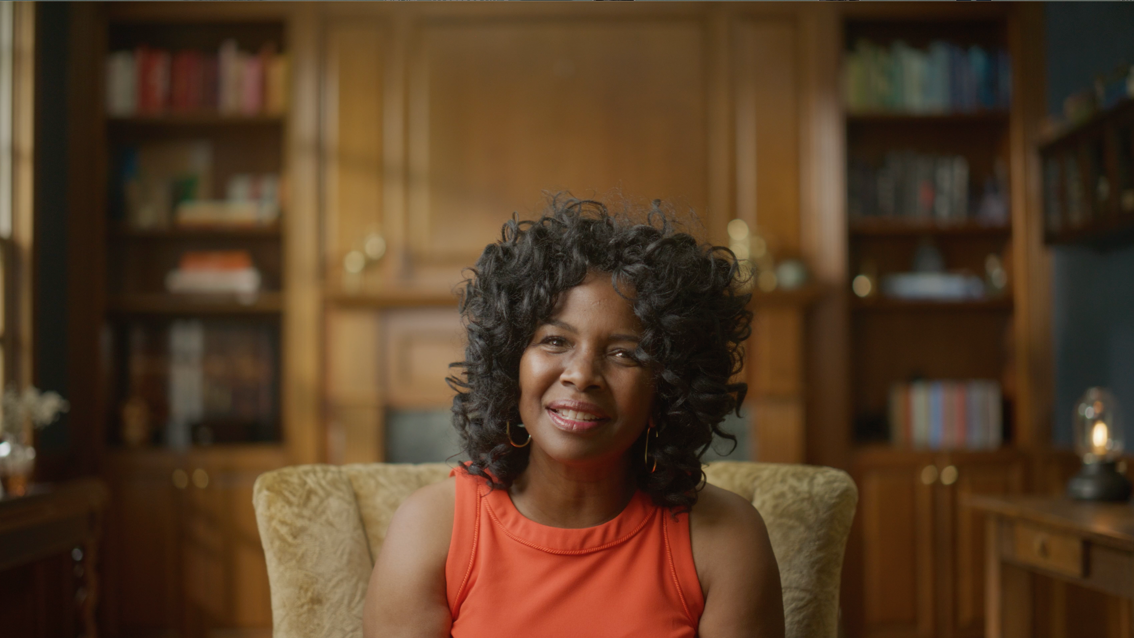 A woman with curly black hair, wearing an orange sleeveless top, sitting in a room with wooden bookshelves filled with books and decorative items in the background.