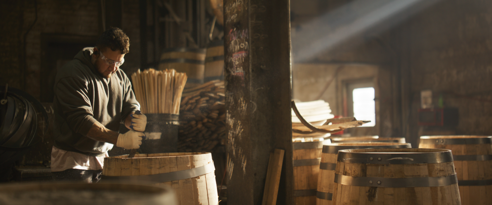 A man working inside a rustic workshop with wooden barrels, handling a woodworking process with sunlight streaming through a window.