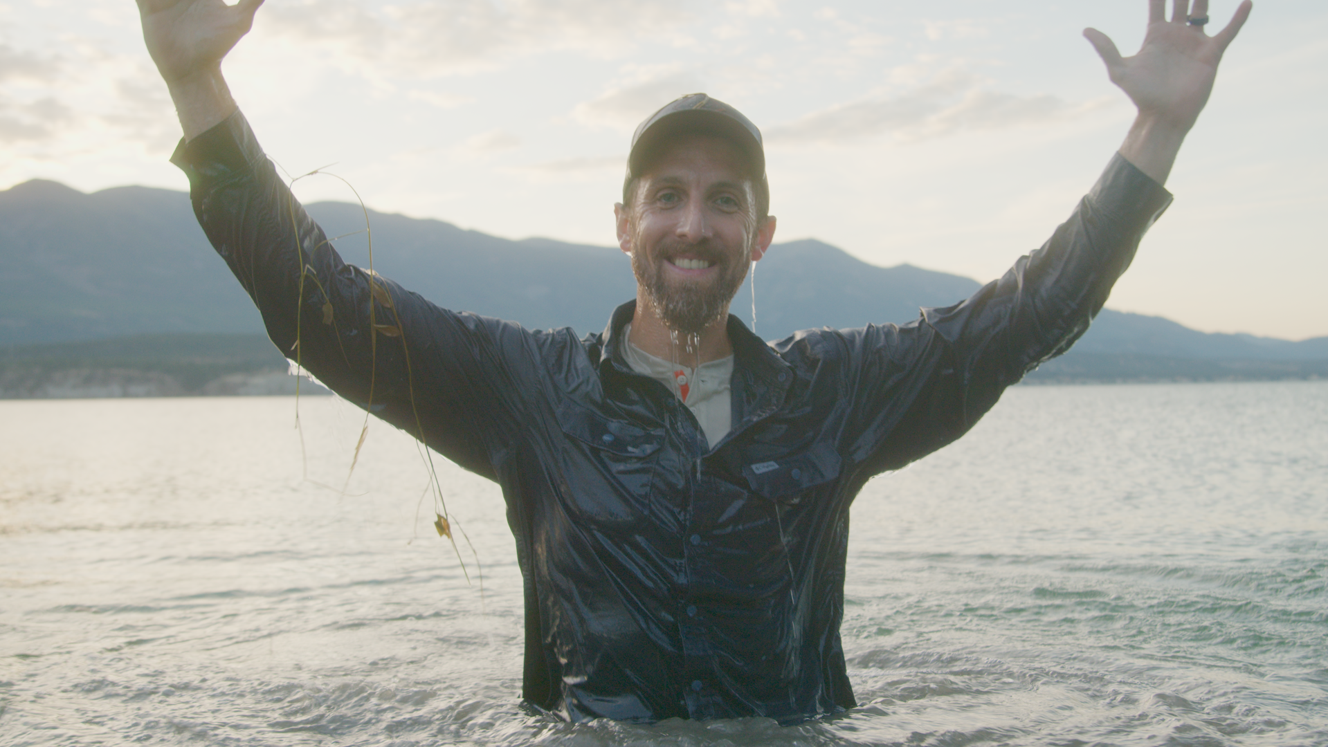 Man standing in water with arms raised, smiling, mountains and cloudy sky in background.