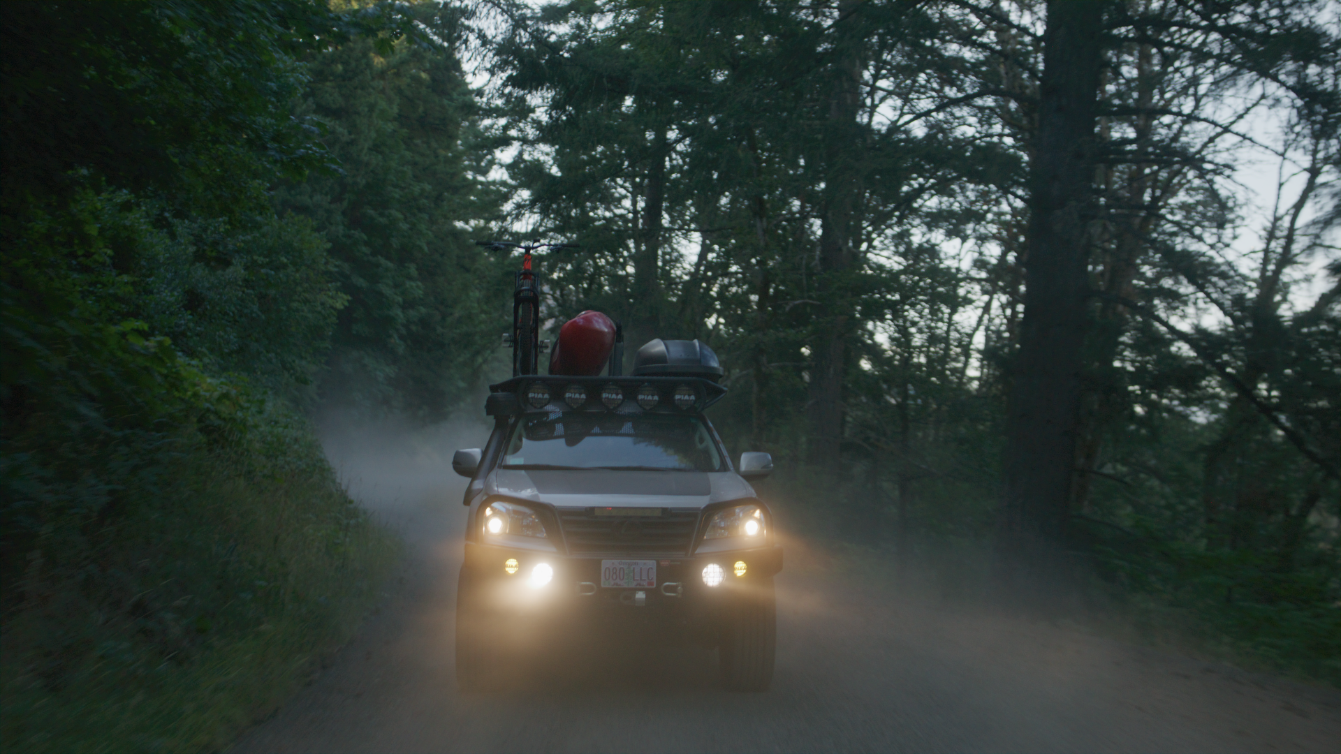 A silver SUV driving through a forest dirt road at dusk with its headlights on, carrying a bike and other gear on the roof rack.