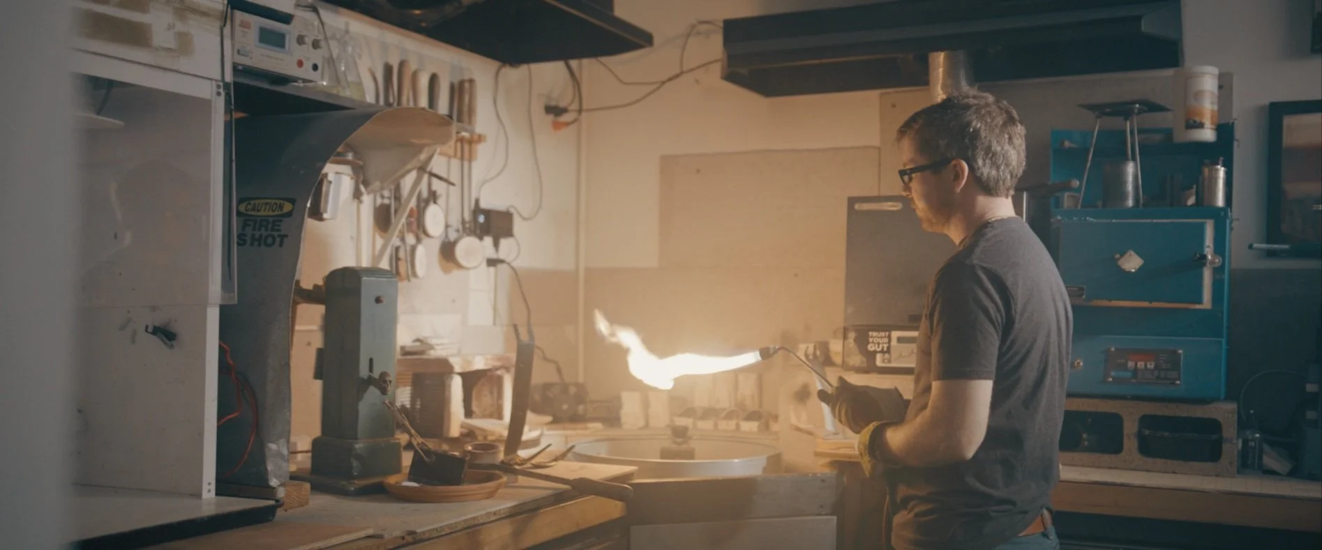 A man in a workshop is holding a torch with a flame, working on a metal piece, surrounded by tools and equipment.