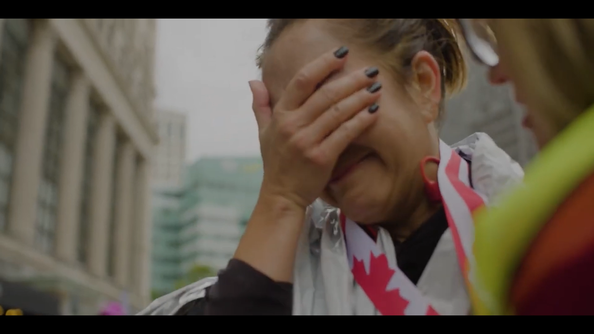 A woman with short hair covering her face with her hand, appearing emotional, during an outdoor event in an urban setting.