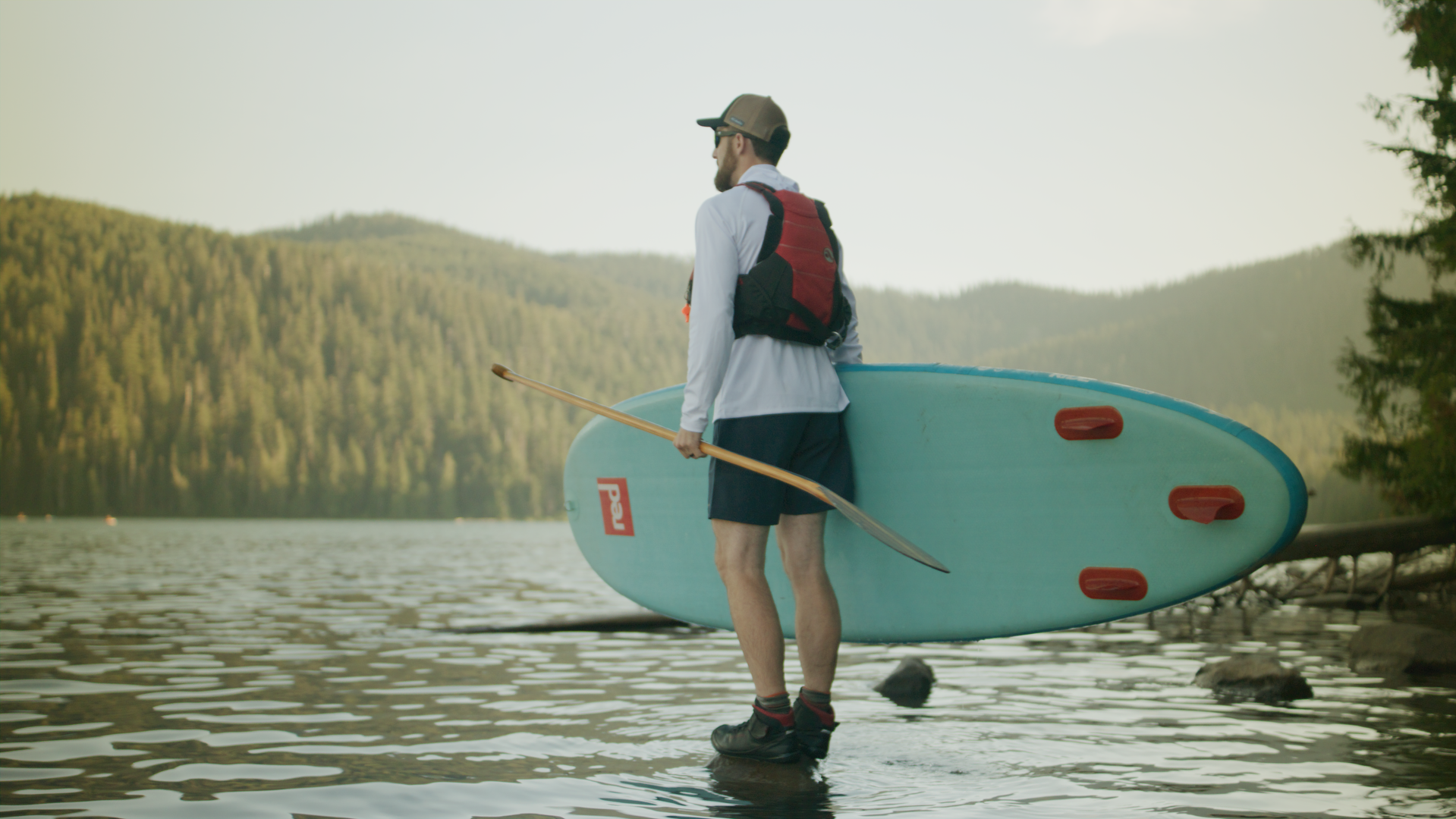 A man standing in shallow water holding a paddle and a paddleboard, near a forested lake shore with mountains in the background.