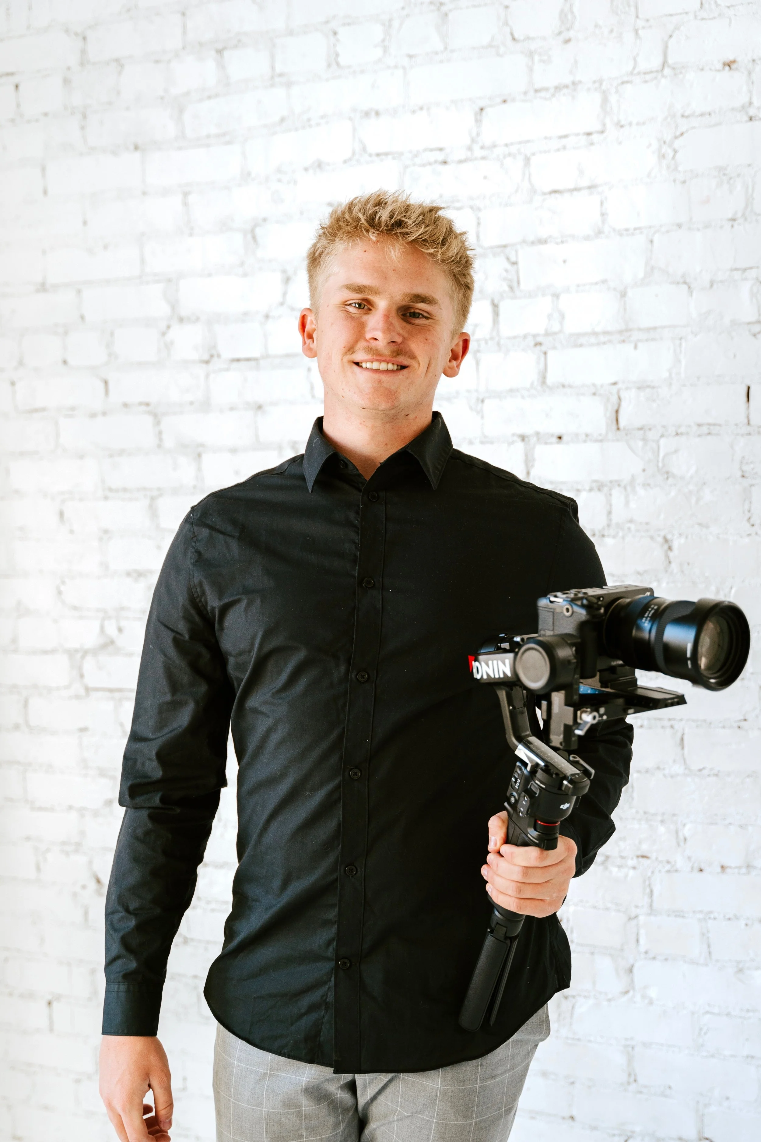 Videographer standing against a white brick wall with camera.