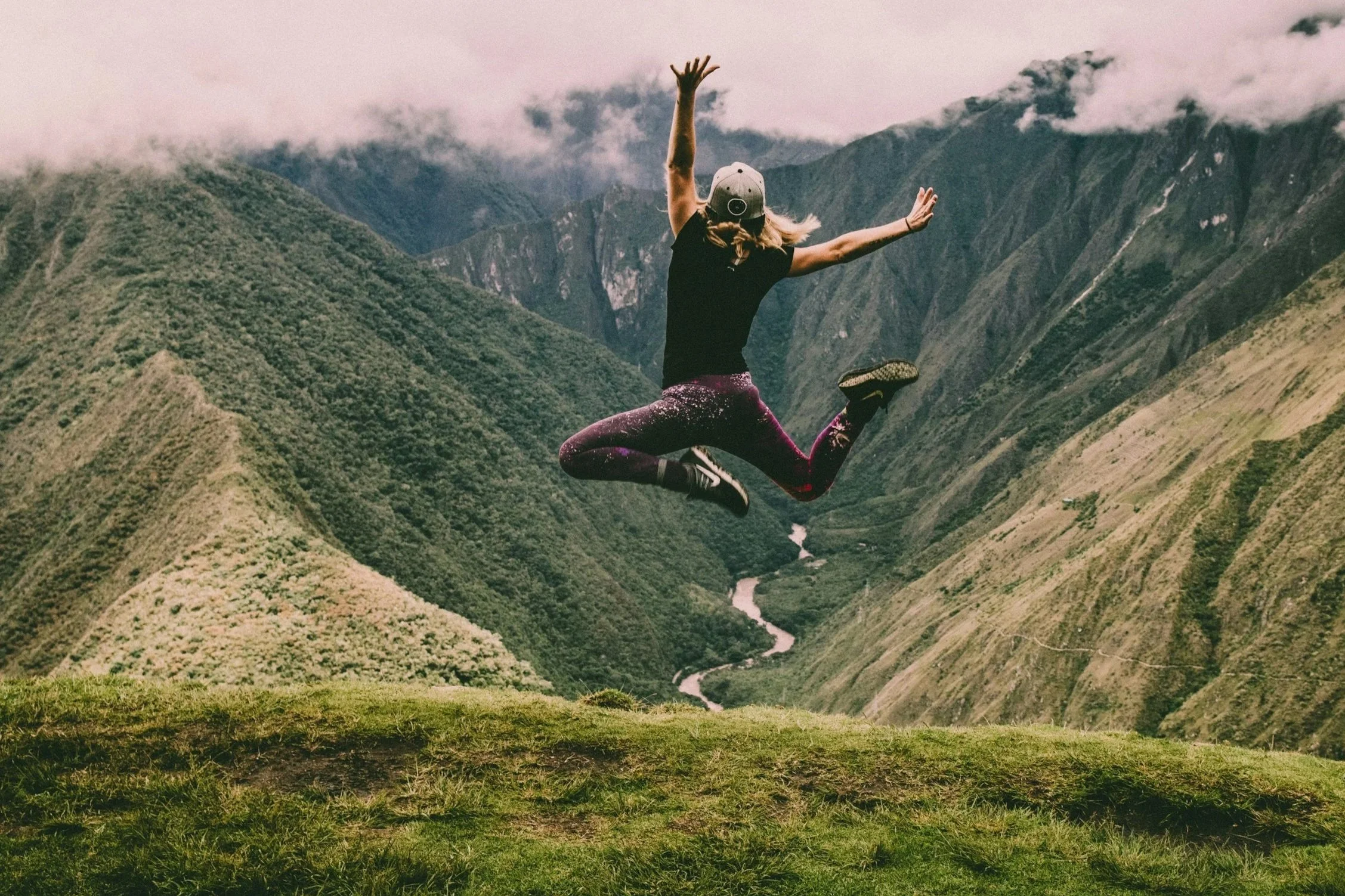 A woman in athletic clothing jumping in the air on a grassy hilltop with a scenic mountain valley and river in the background.