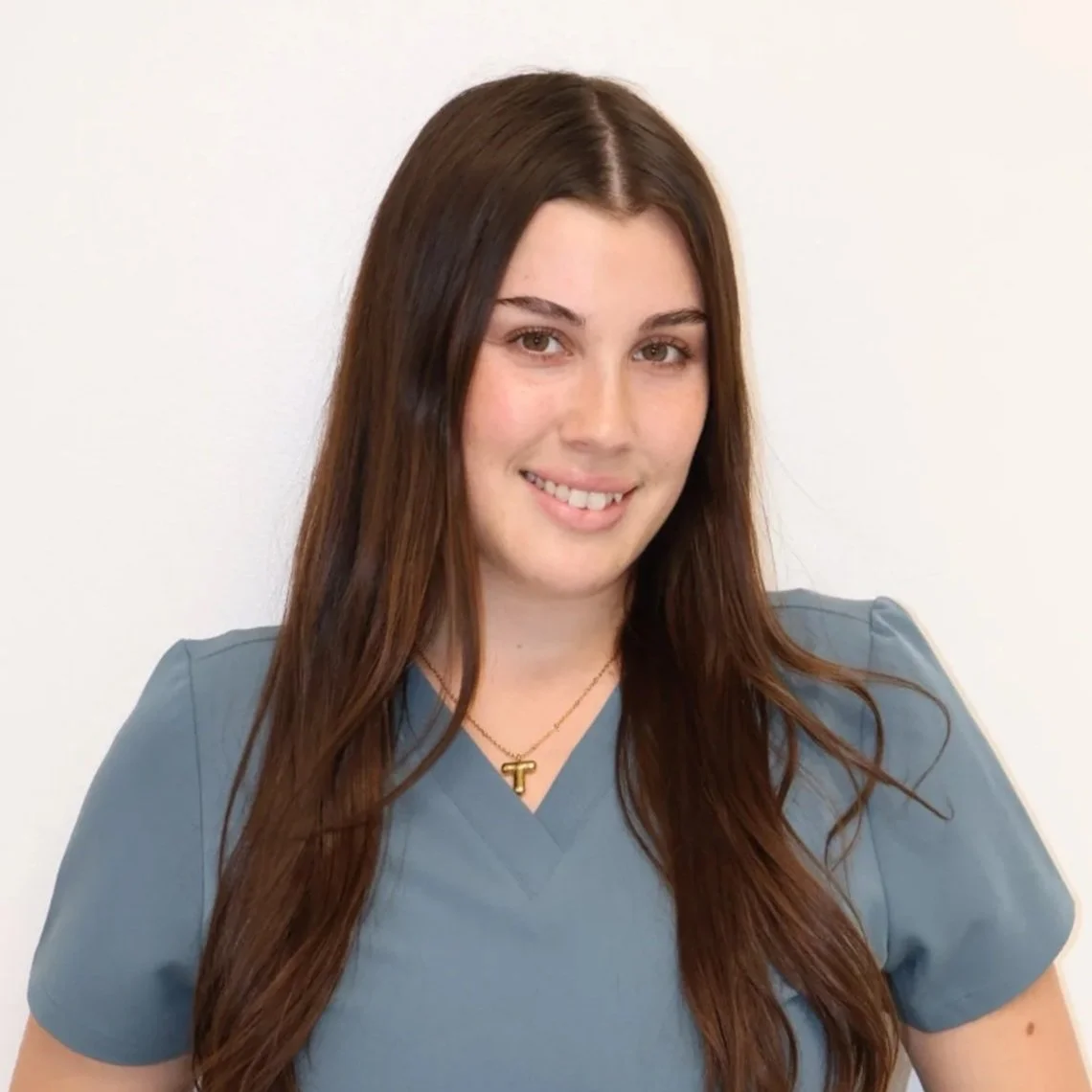 Portrait of a woman with long dark hair, smiling, wearing an off-the-shoulder green top and a delicate necklace, against a plain background.