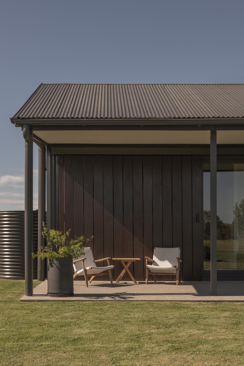 A modern house patio with two chairs, a small wooden table, a large potted plant, and a corrugated metal roof overhang, surrounded by green grass and clear sky.