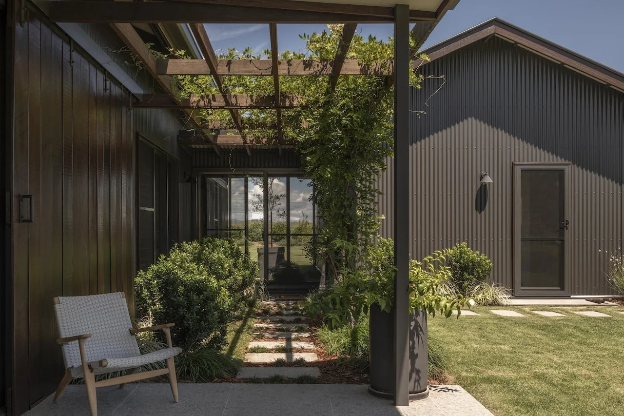 Exterior view of a modern backyard with a shaded patio area, lush green plants, a small wooden bench, and a pathway leading to a glass door.