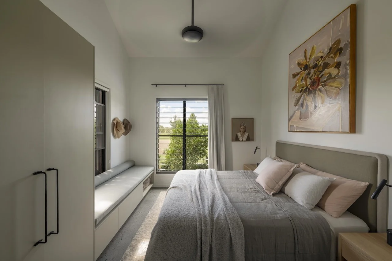 A cozy bedroom with a large bed, pastel pillows, artwork on the wall, a window with curtains, and minimal decor.