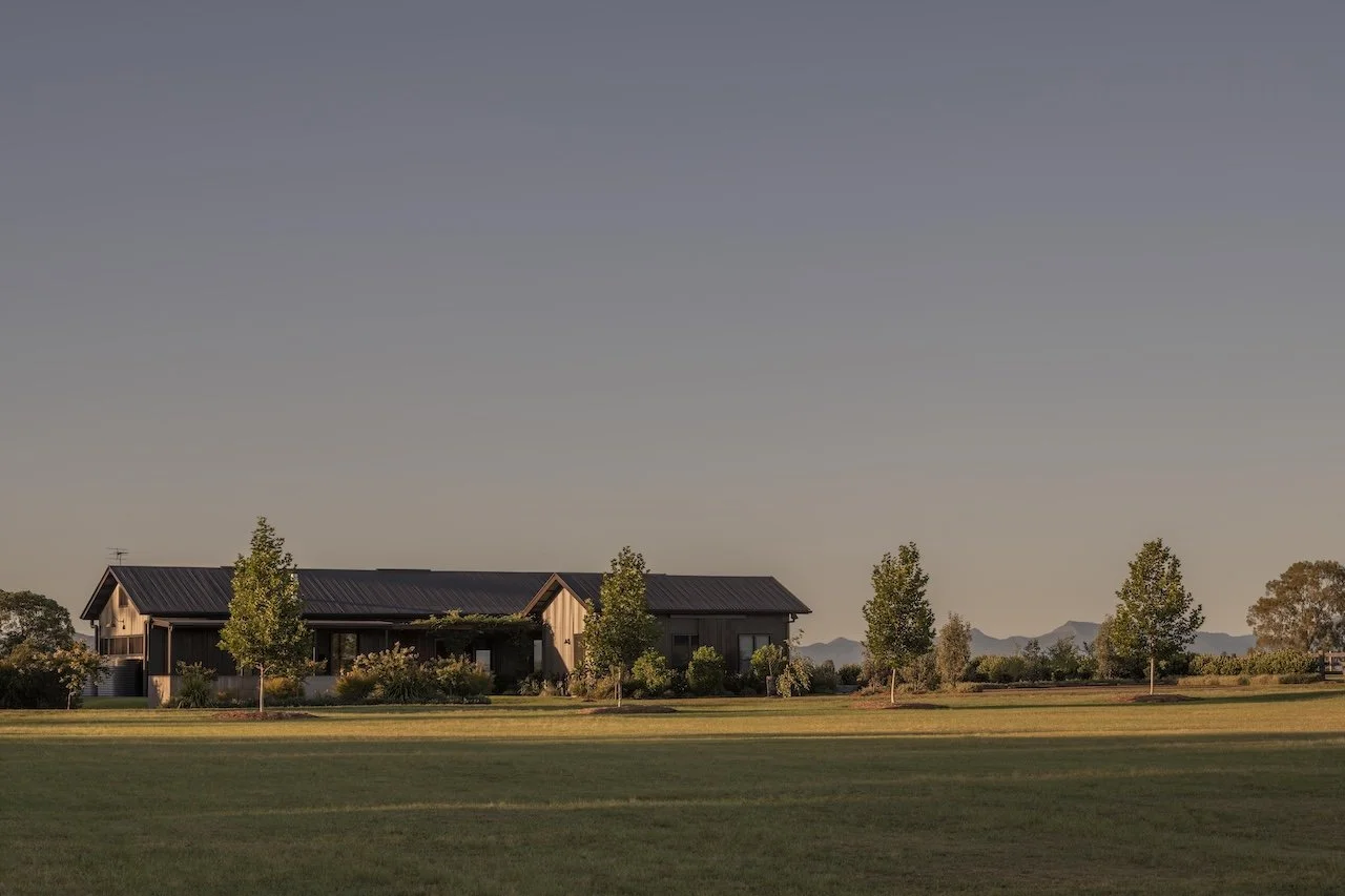A house with a metal roof surrounded by trees and a large green lawn, with distant mountains in the background under a clear sky.
