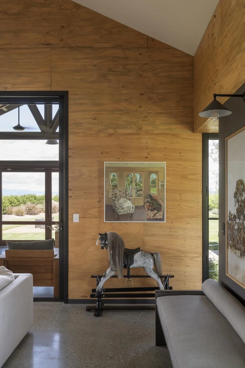 Interior of a room with wood-paneled walls, a rocking horse with a saddle, a painting of an interior scene, and large windows showing a yard and outdoor landscape.