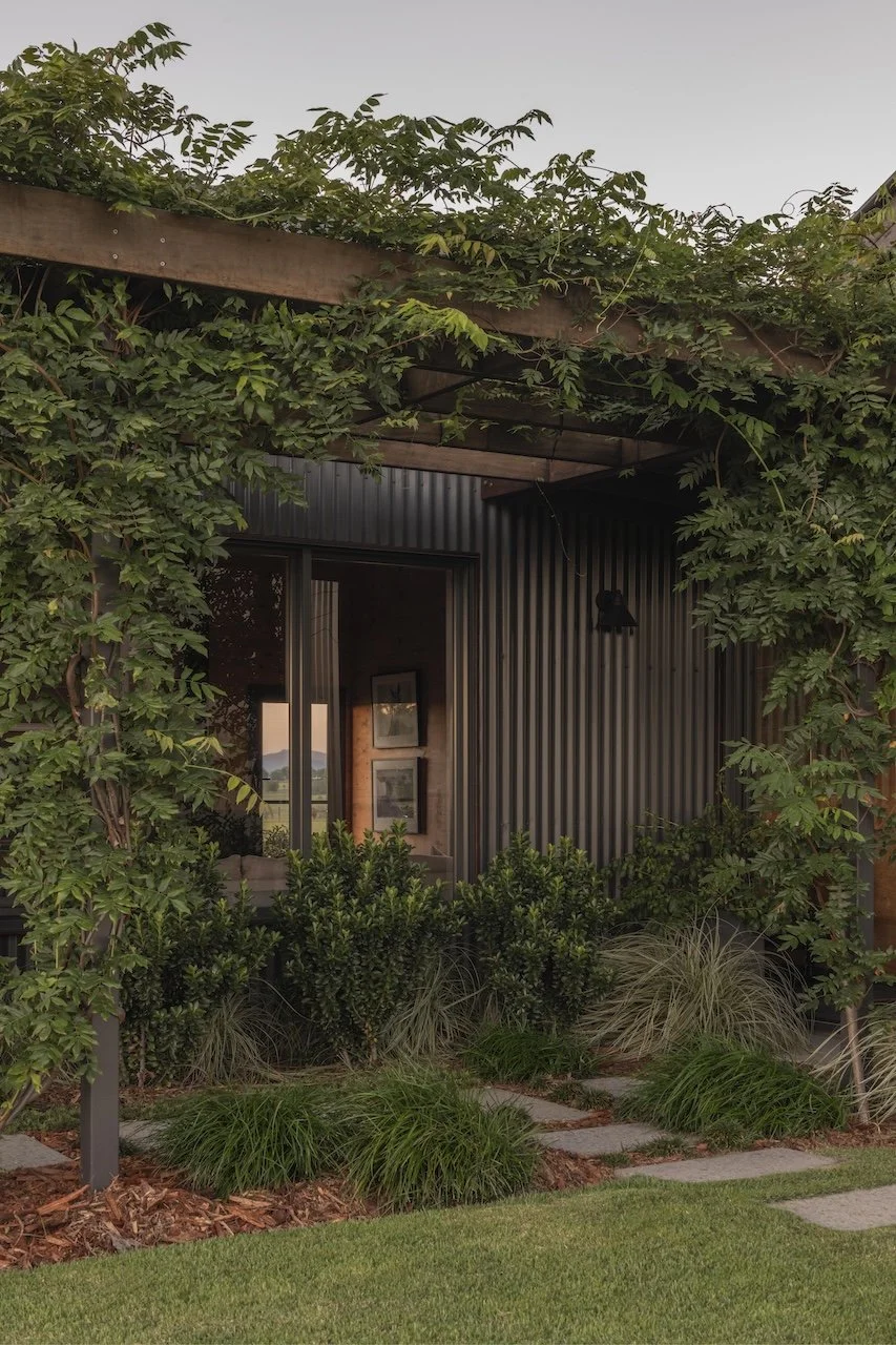 Exterior view of a house with metal siding, surrounded by lush green plants and trees, at dusk.