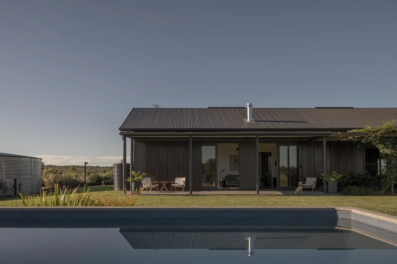 Modern black house with a patio, outdoor furniture, trees, and a swimming pool in the foreground under a clear sky.
