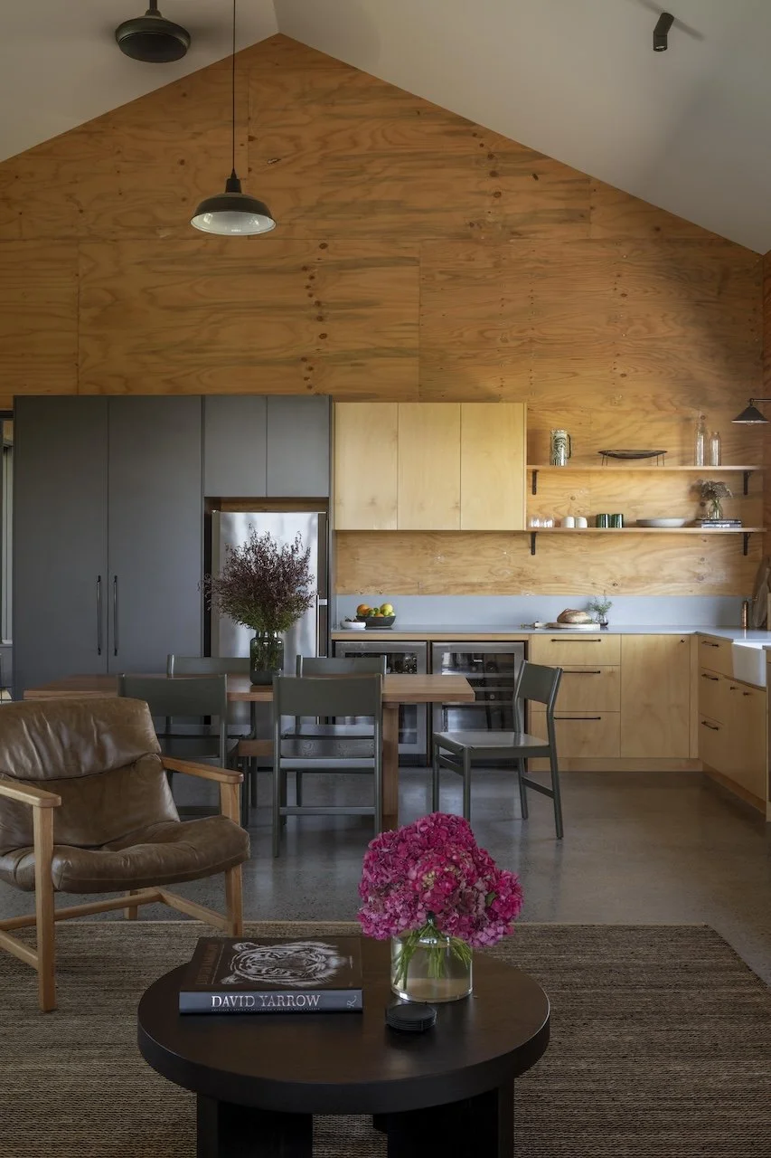 Modern kitchen and living space with wood-paneled walls and ceiling, gray and wooden cabinets, a dining table with chairs, a brown leather armchair, a dark wood coffee table with a pink flower arrangement and a book, and two hanging black pendant lig