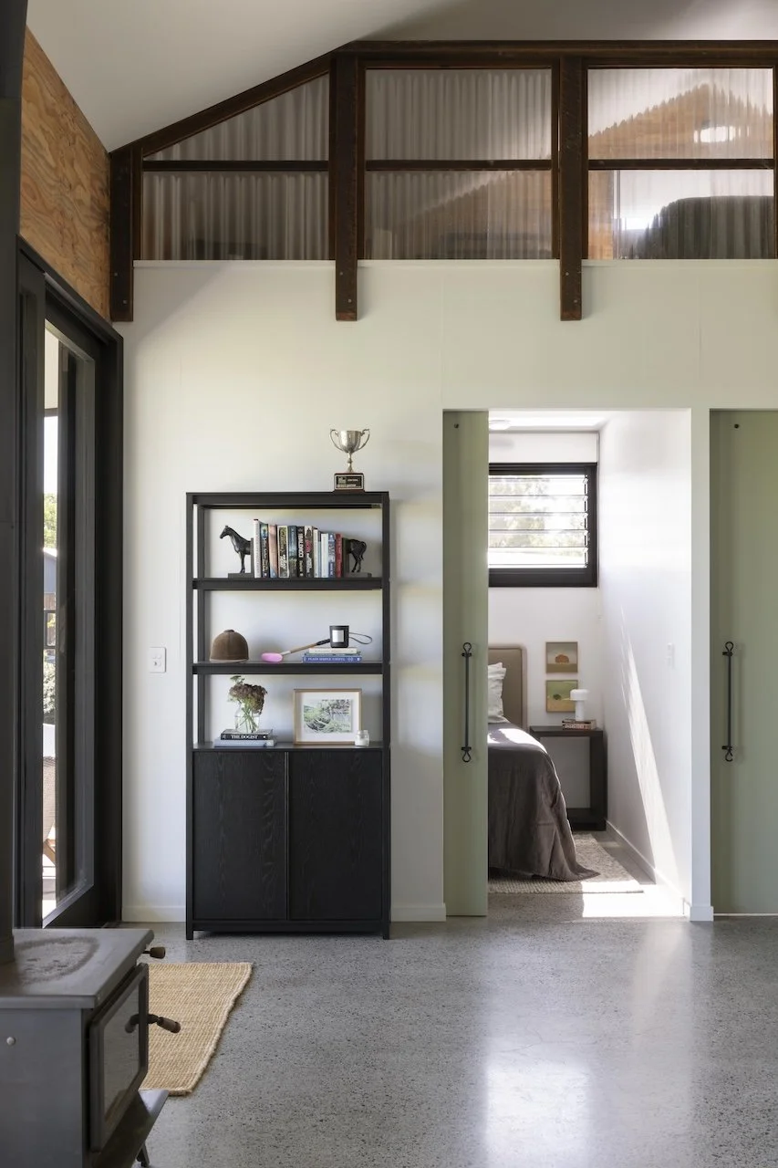 A corner of a modern room with a black shelving unit displaying books, a trophy, and decorative items; a small bedroom visible through an open doorway with a bed and nightstand; and a large window with blinds in the background.
