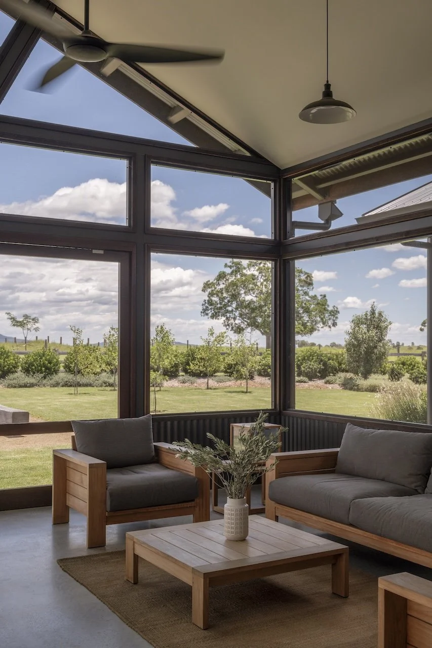 A sunlit living room with large glass windows showing a backyard landscape with trees and a cloudy sky. Inside, there are wooden furniture pieces, including two chairs and a coffee table with a vase of greenery, all on a beige rug. A ceiling fan and a hanging light fixture are also visible.