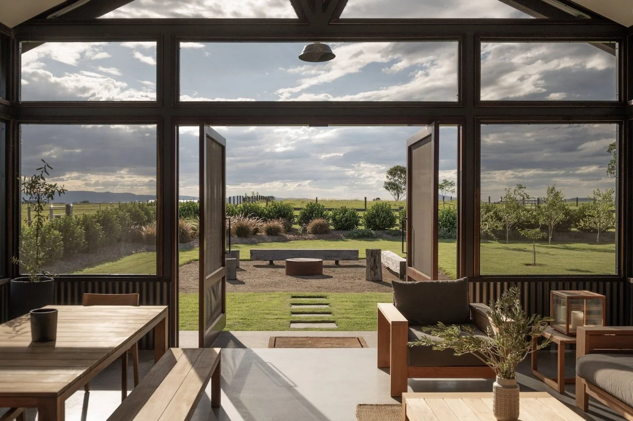 Interior of a modern living room with large open glass doors leading to a garden view with grass, shrubs, and trees. The room contains wooden furniture including a table, benches, and a sofa with cushions. There are decorative plants and a candle on 