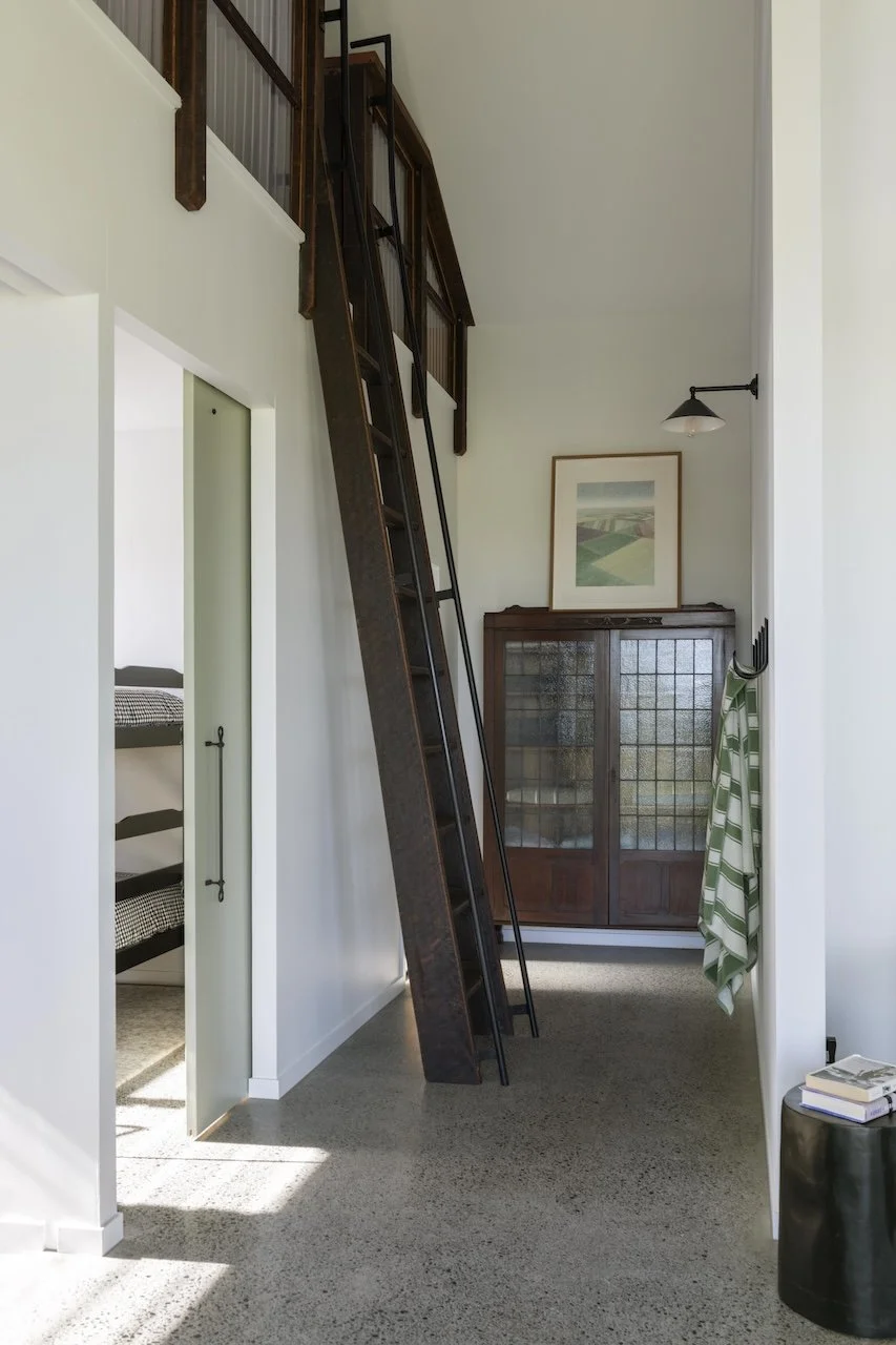 Interior view of a room with a wooden loft ladder leading to a small upper storage or sleeping space. There is a wooden cabinet with glass doors at the end of the room, a framed picture above it, a wall-mounted light, a green and white striped towel 