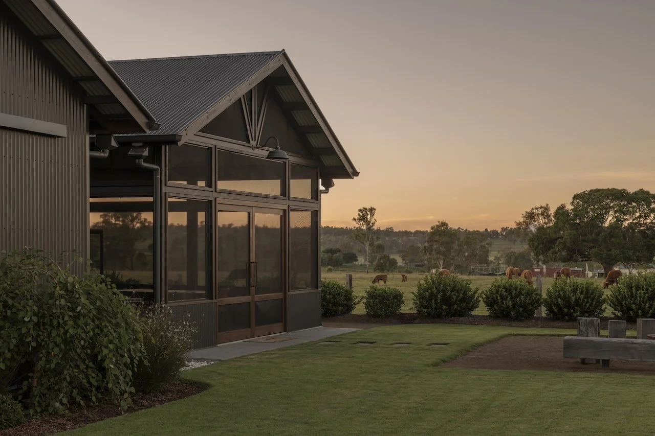 A modern building with large glass windows and a metal roof, overlooking a rural landscape with horses grazing at sunset.