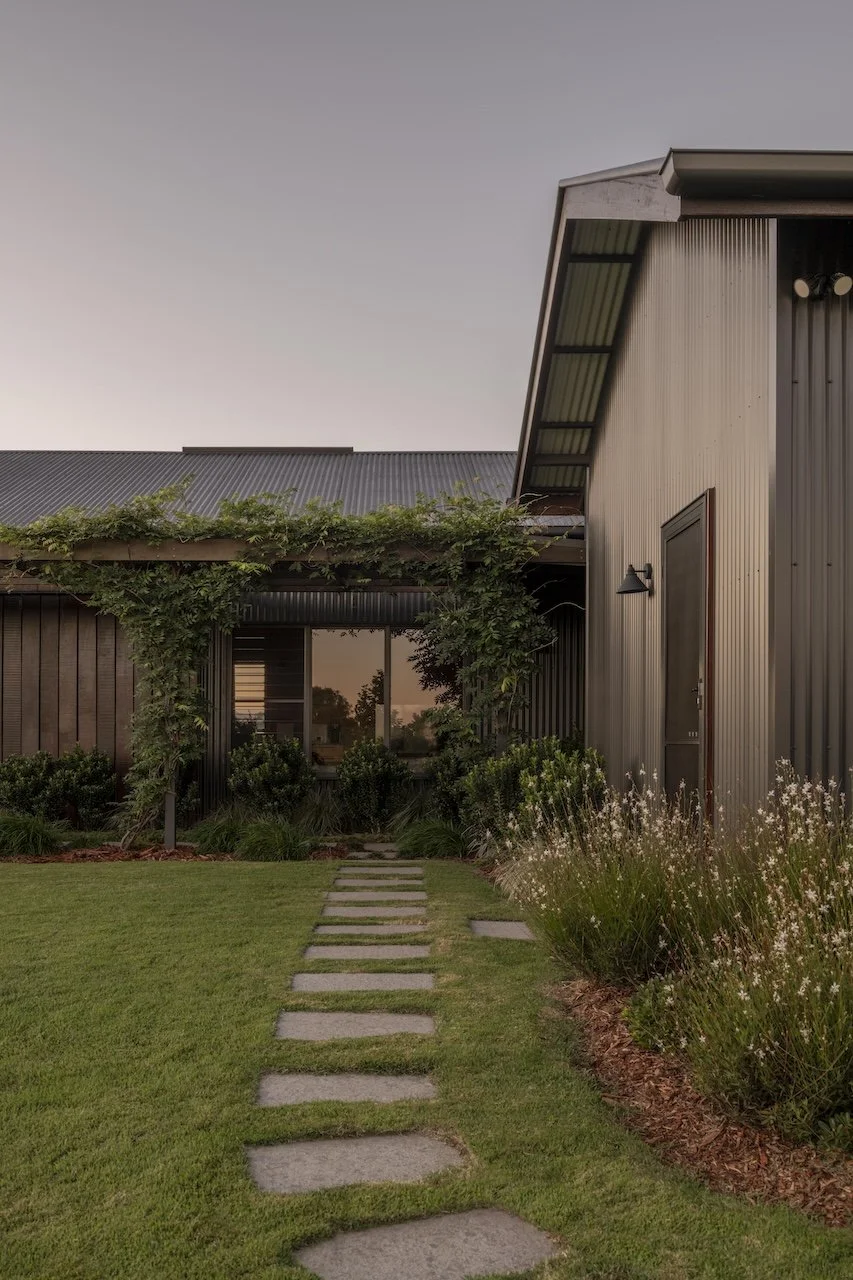 Front yard of a modern house with a stone pathway, green grass, bushes, flowering plants, and a house with metal siding and large windows at sunset.