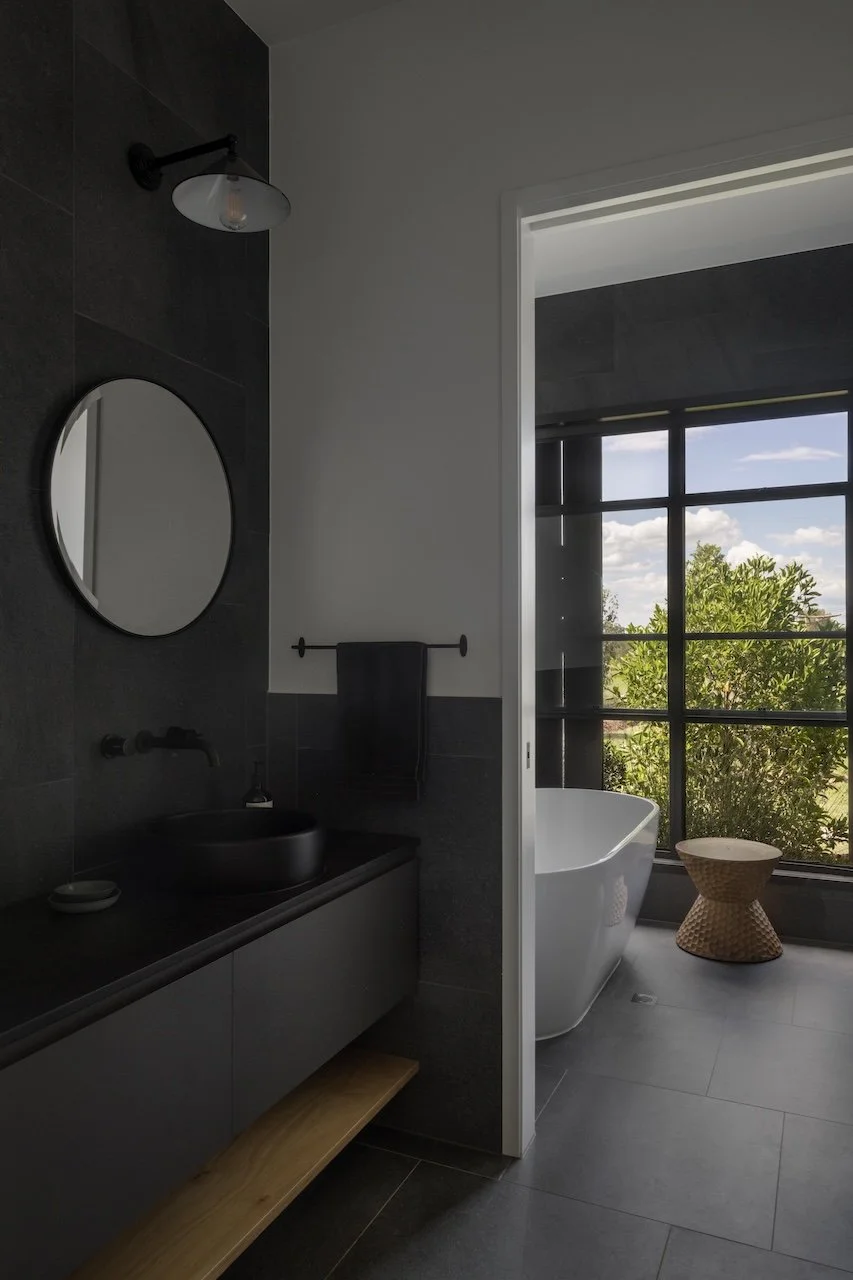 Modern bathroom with dark vanity, round mirror, black vessel sink, black towel, and a large window showing greenery outside and a freestanding white bathtub.