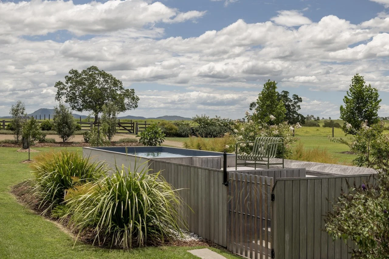 A backyard with a small above-ground pool, a metal bench on top, surrounded by green grass, various plants, trees, and a wooden fence in the distance under a partly cloudy sky.