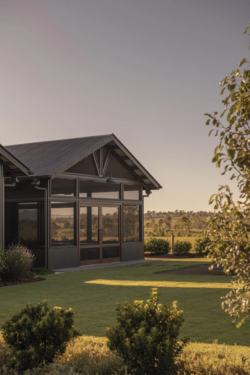 A modern black house with large windows and a metal roof, situated in a green lawn with bushes, during sunset or early evening.