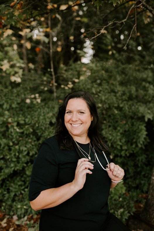 A woman with dark hair smiling outdoors, holding a pair of glasses, standing in front of green bushes and trees.