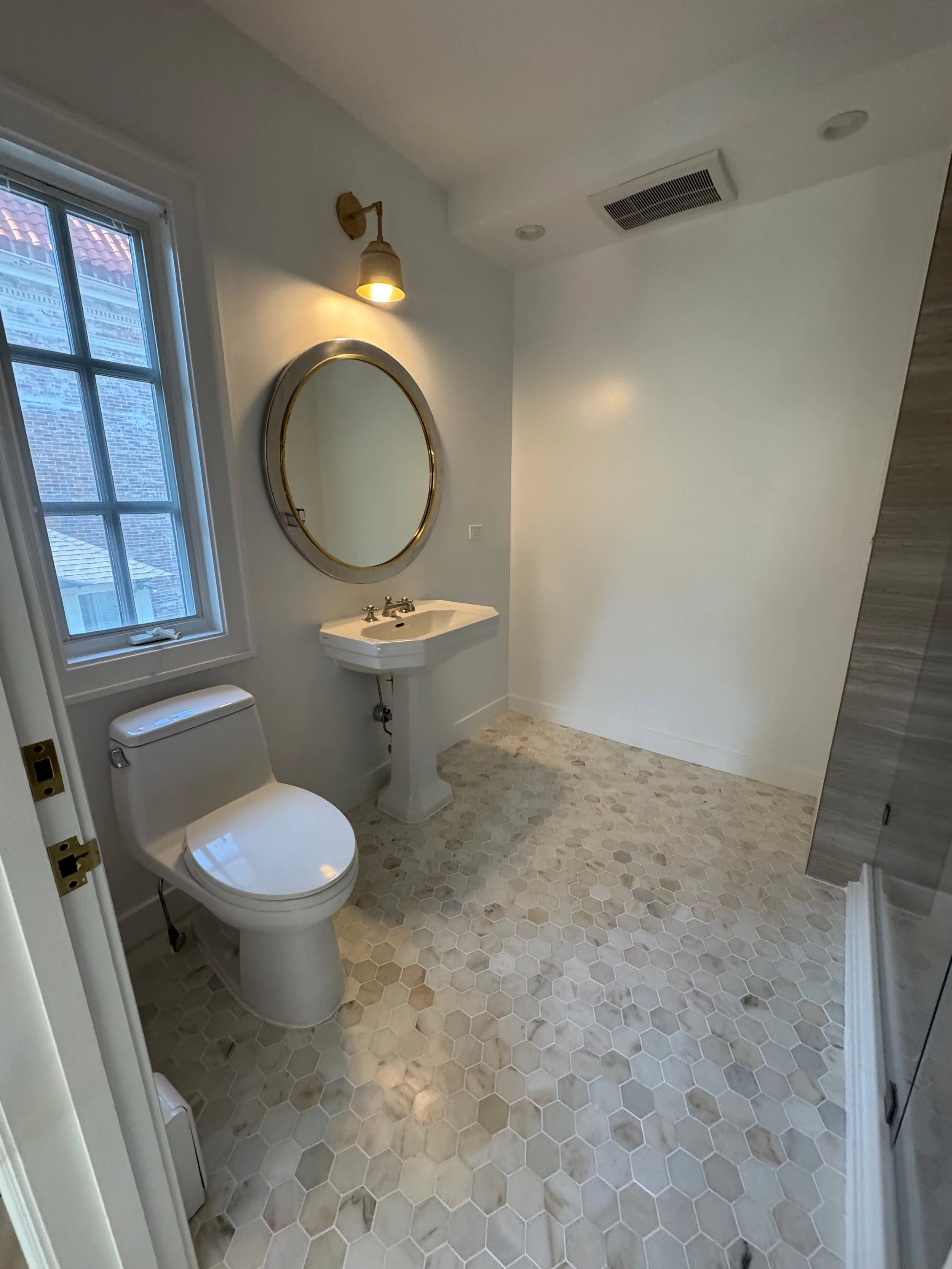 A small bathroom with a white toilet, a small white pedestal sink, a round gold-framed mirror, a wall-mounted light fixture, a window with a view of neighboring houses, and a hexagonal tile floor.