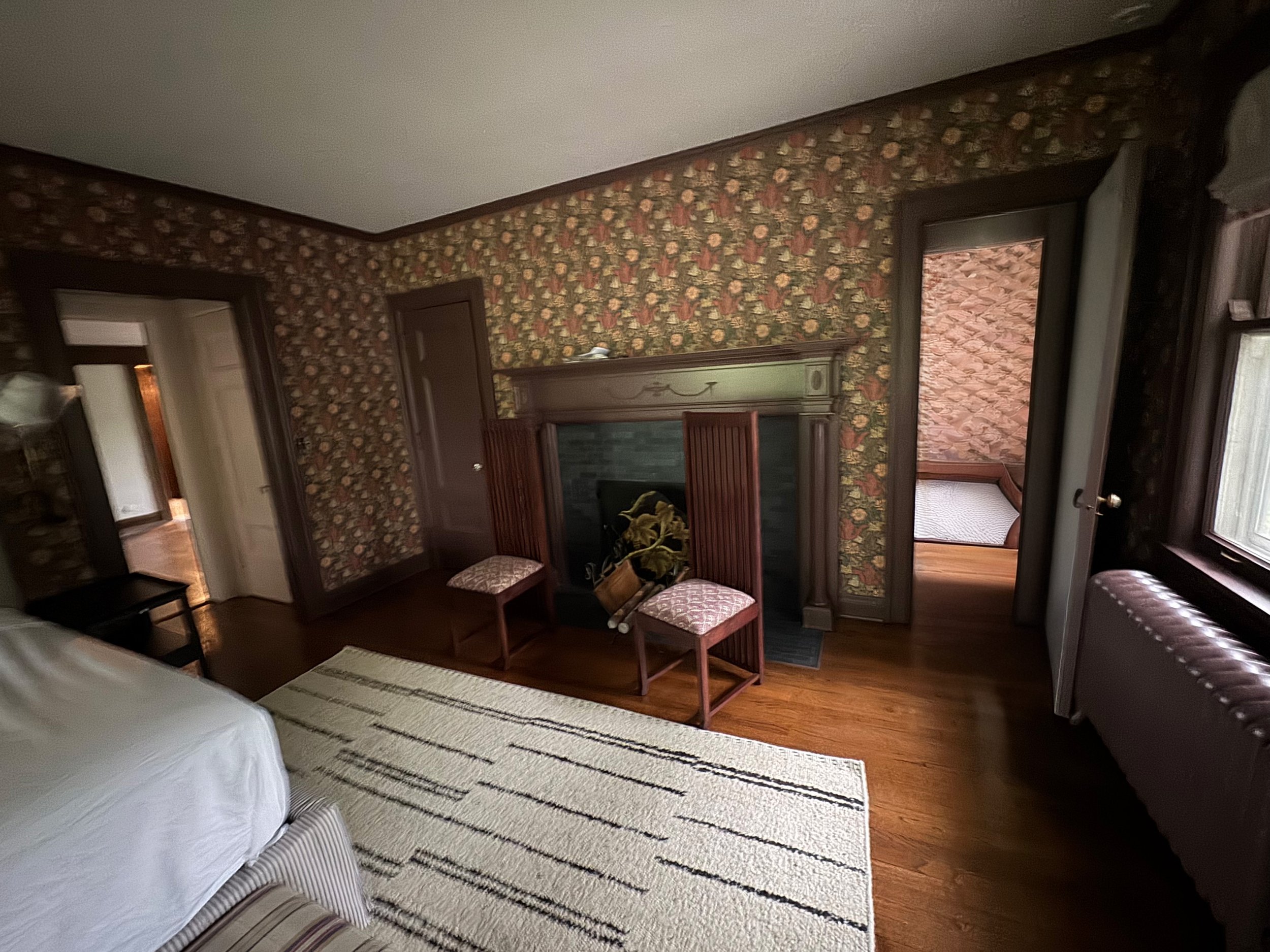 A cozy bedroom with floral wallpaper, hardwood floors, a bed with white linens, a fireplace with two wooden chairs, a window with birch bark decor in the adjacent room, and a patterned area rug.