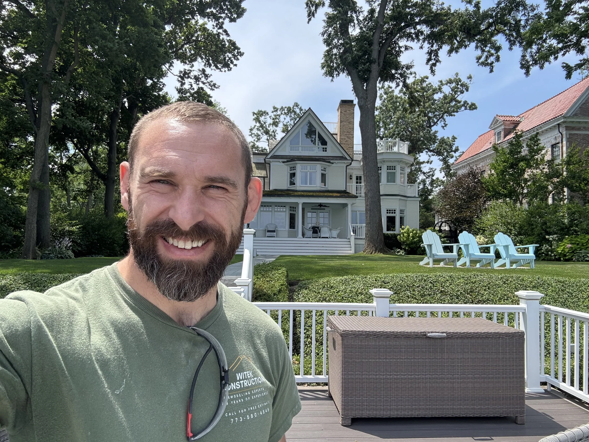 A smiling man with a beard taking a selfie on a backyard deck with a white house featuring a large porch and multiple balconies, surrounded by trees and outdoor furniture including white Adirondack chairs.