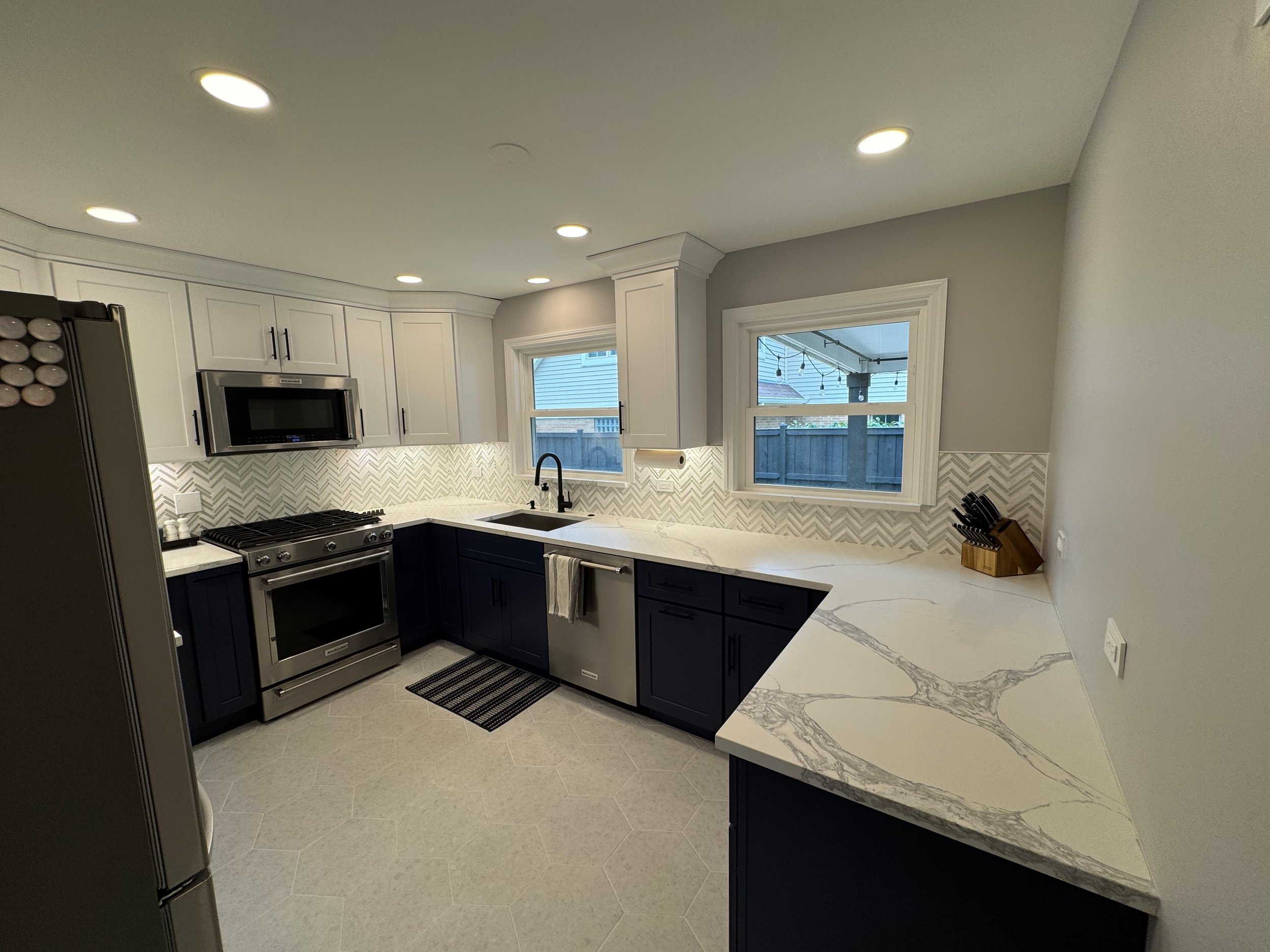 Modern kitchen with white upper cabinets, navy lower cabinets, white marble countertops, a stainless steel stove and microwave, a black sink, and two windows overlooking a backyard.