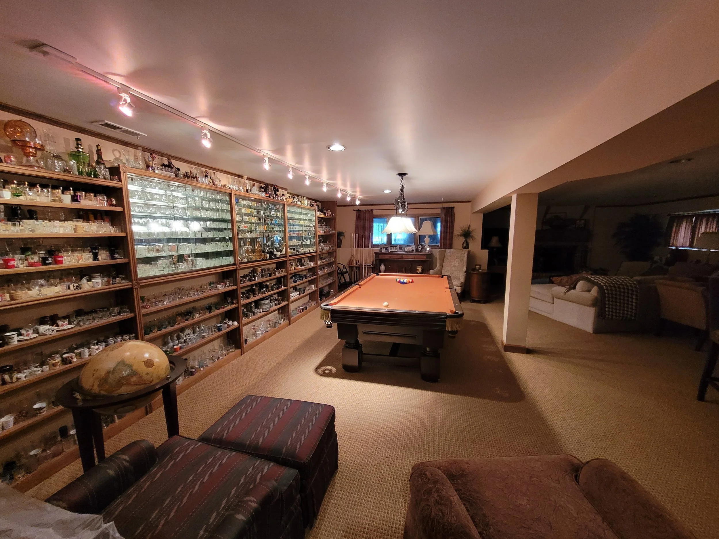 A cozy basement with a pool table, surrounded by shelves filled with glass collectibles, and a seating area with a sofa and chairs, illuminated by warm lighting.