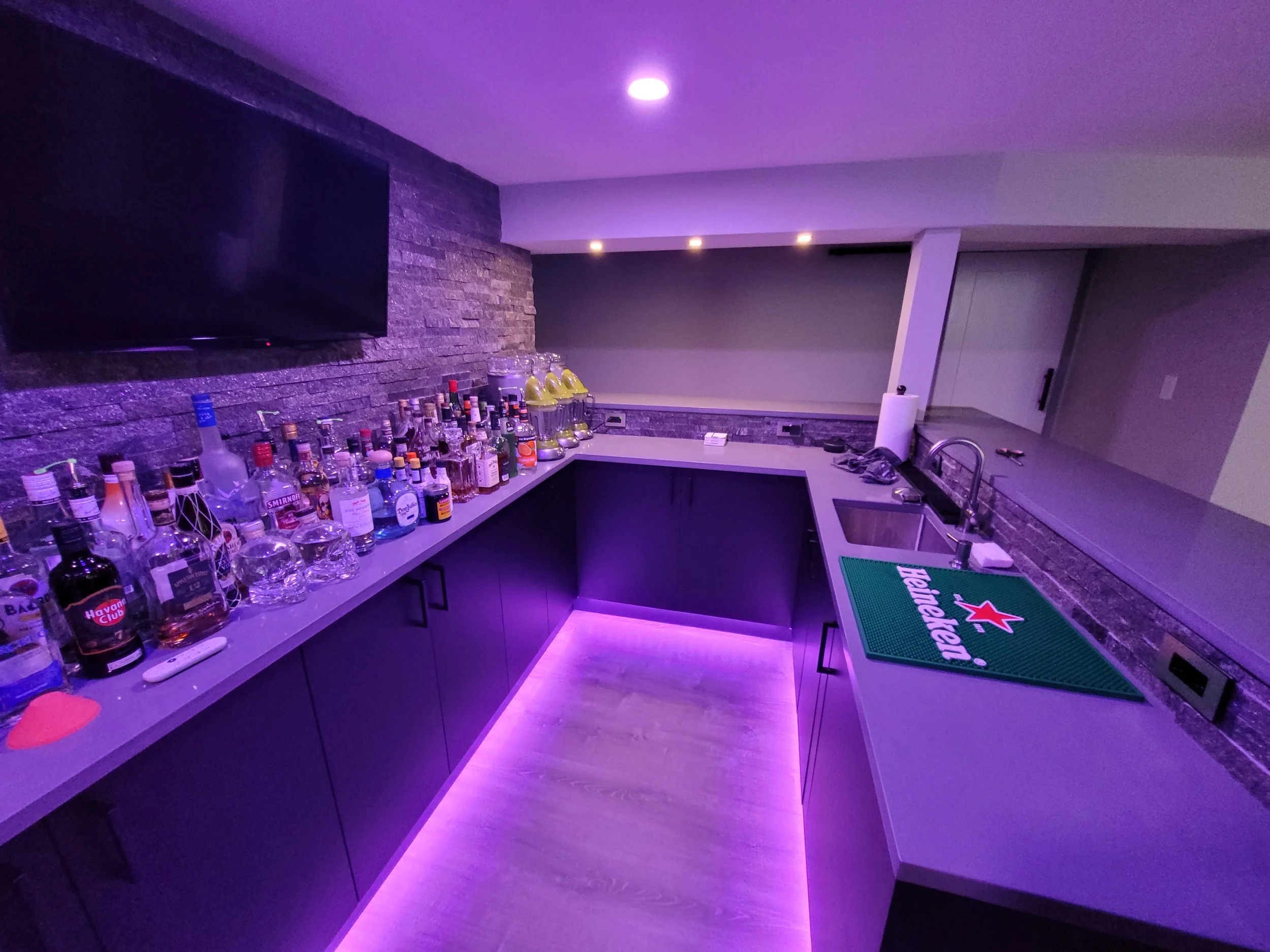 A modern home bar with a purple lighting accent, a large television mounted on a brick accent wall, a countertop filled with various bottles of alcohol, a Heineken bar mat at the sink, and minimalistic cabinetry.