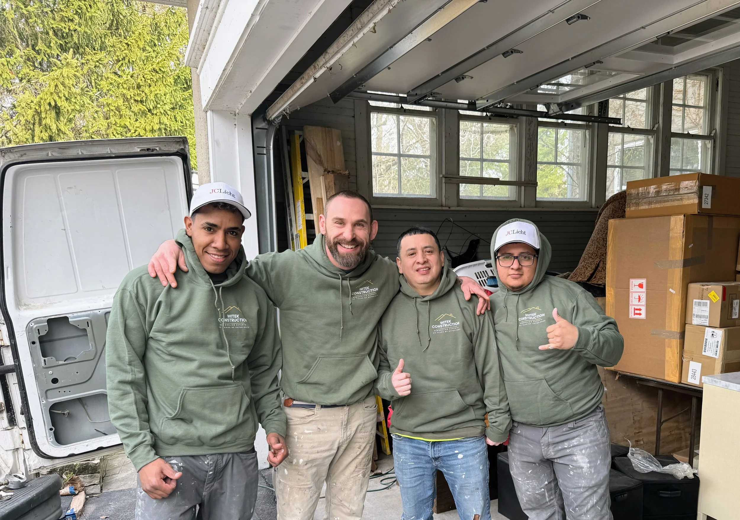 Four men in matching green hoodies and two wearing white caps, standing in a garage with boxes and tools, smiling for the photo.