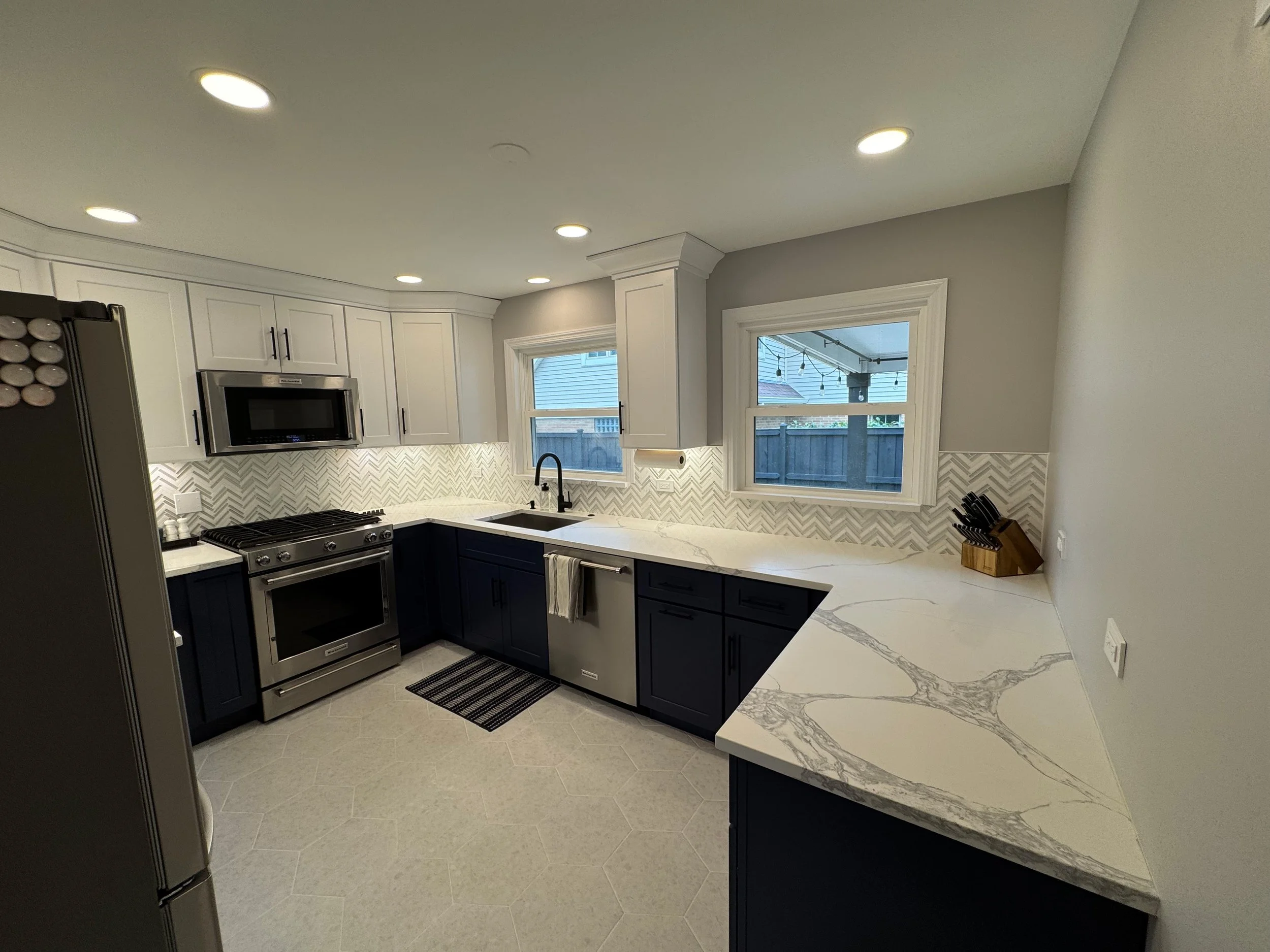 Kitchen with white upper cabinets, navy blue lower cabinets, stainless steel appliances, marble countertop, and light gray walls.