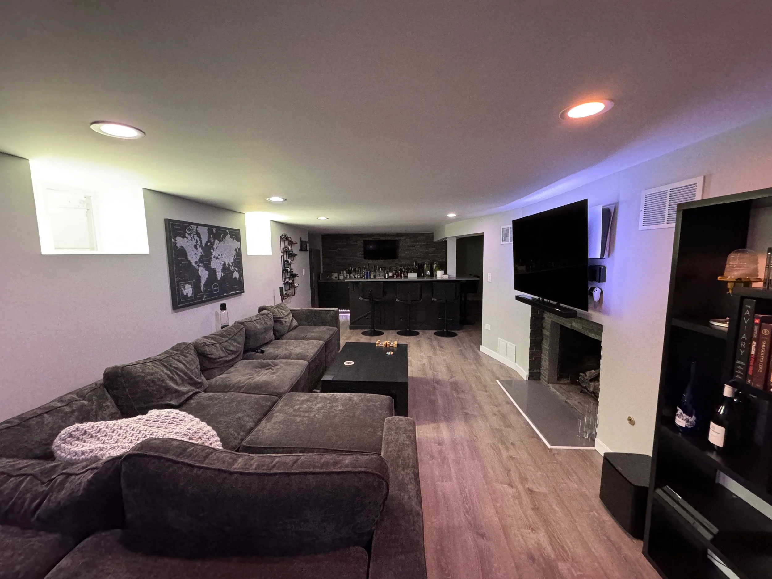 A cozy basement living room with a large gray sectional sofa, a black coffee table, and a wall-mounted flat-screen TV above a fireplace. The room features a bar area with stools and a dark accent wall with a world map and wall decor. Recessed lighting and purple-blue accent lighting provide ambiance, with wood flooring throughout.