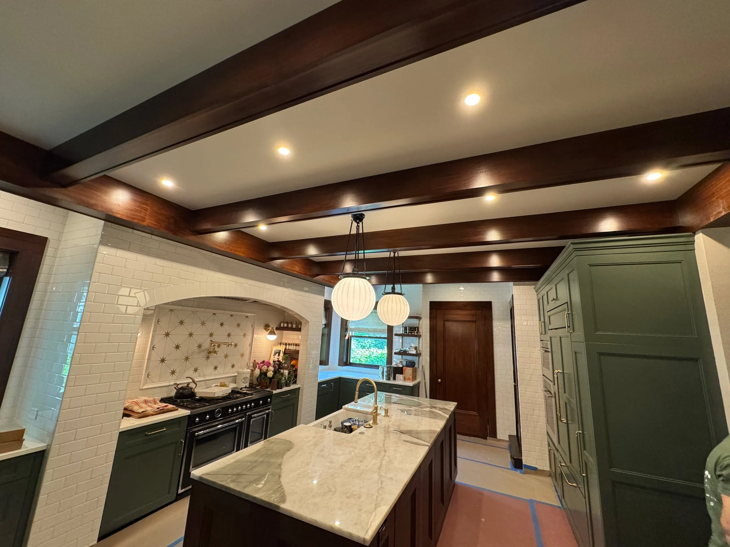 Modern kitchen with dark wood ceiling beams, white subway tile walls, green cabinets, a marble island, and two hanging pendant lights.