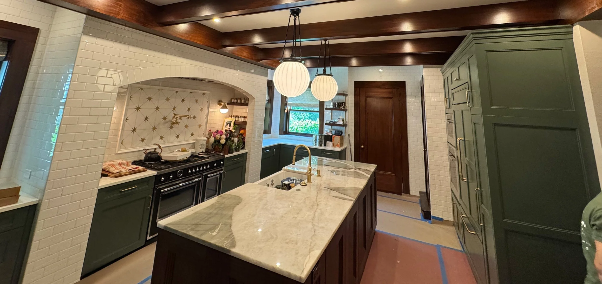 Modern kitchen with green cabinetry, a marble island with a gold faucet, a black range stove, white brick walls, wooden ceiling beams, hanging white globe pendant lights, and a window overlooking greenery.