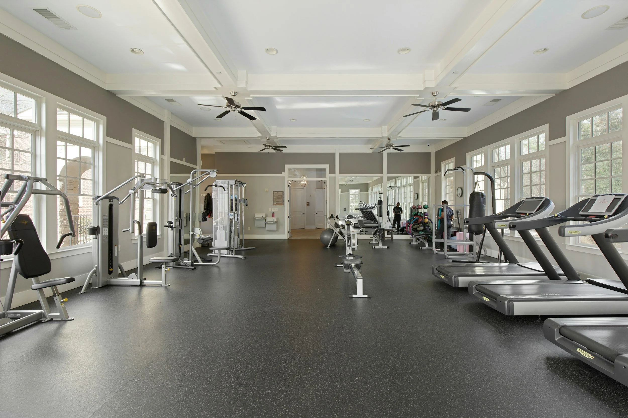 Empty gym with exercise machines, treadmills, weights, and large windows letting in natural light.