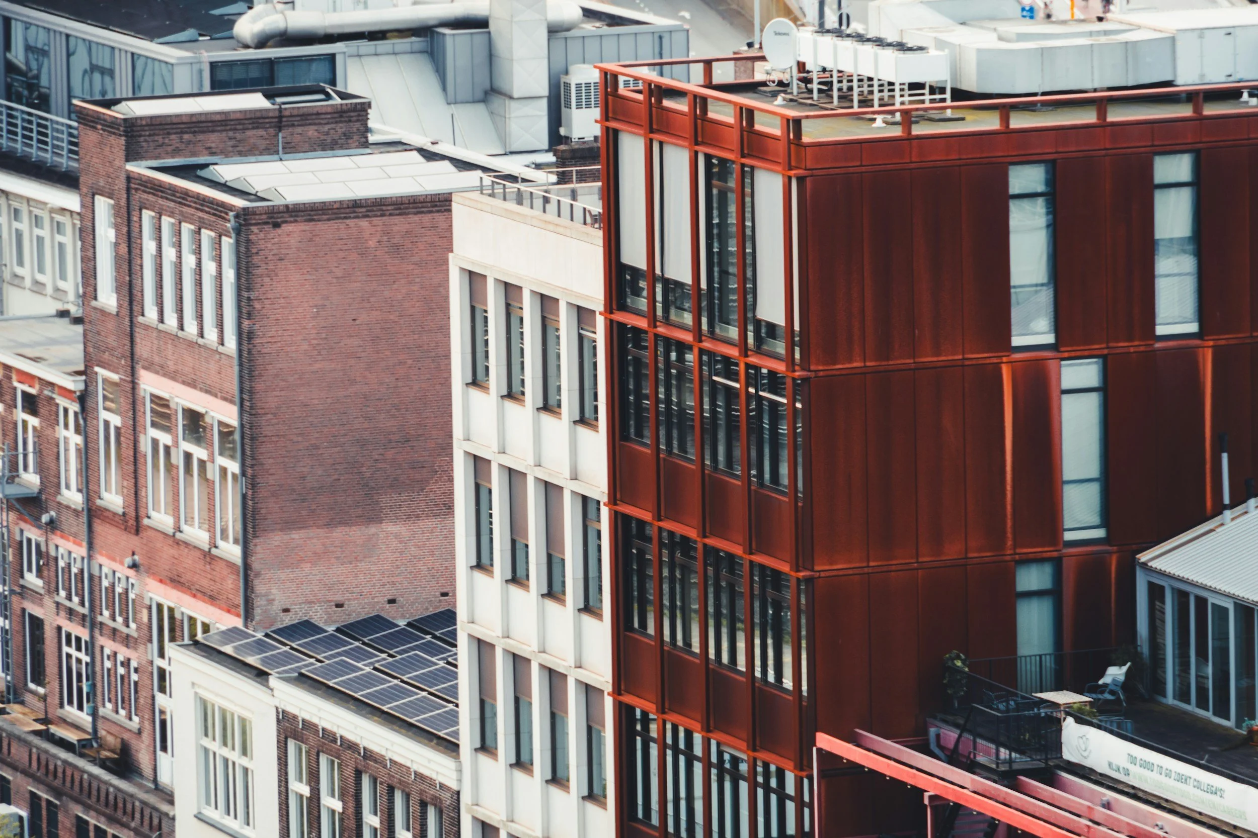 Aerial view of tall urban buildings, including a modern red metal and glass structure, brick buildings, solar panels, and rooftop equipment.