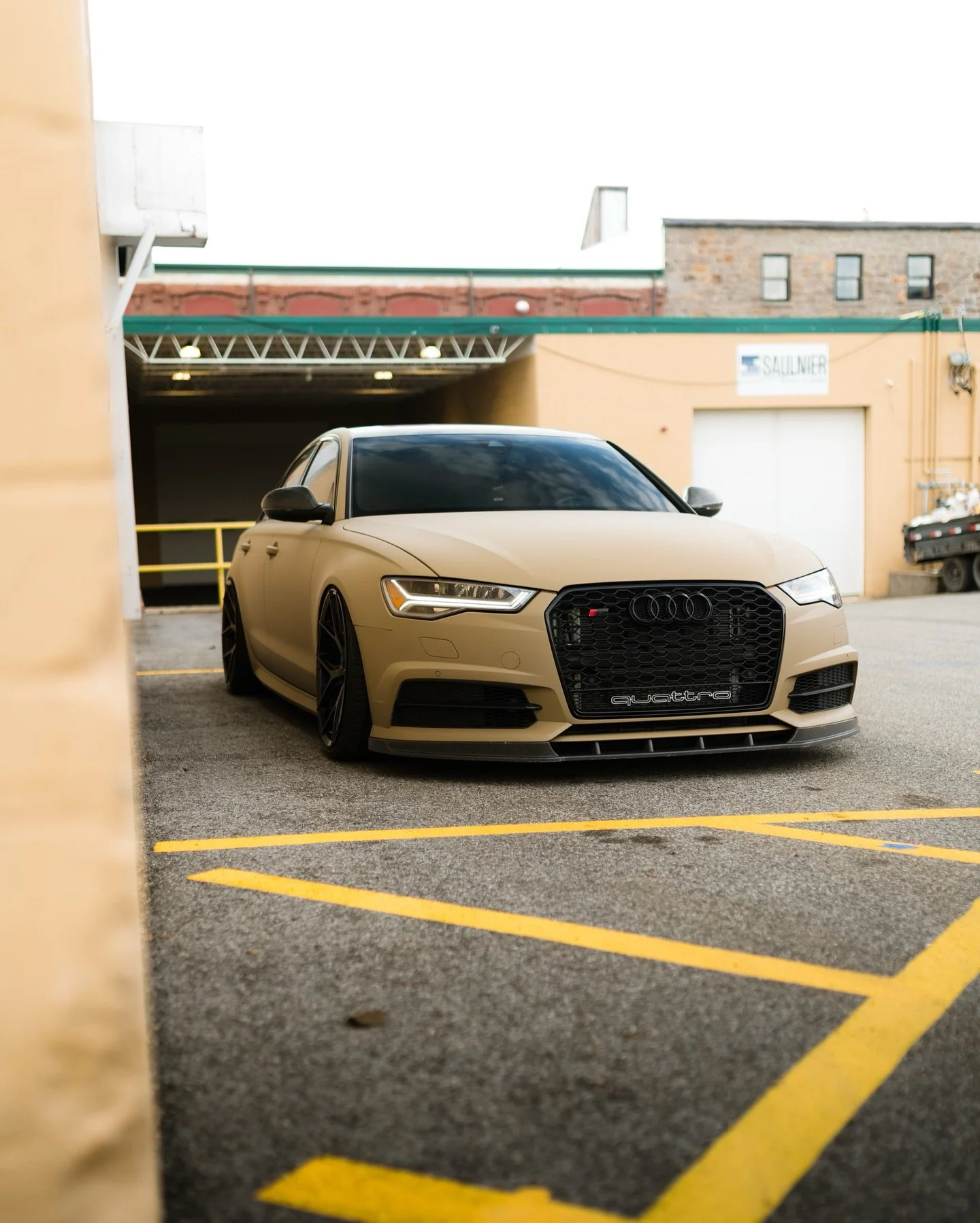 AnAudi RS sedan with a matte beige vinyl wrap parked in a parking lot, with a building and garage in the background.