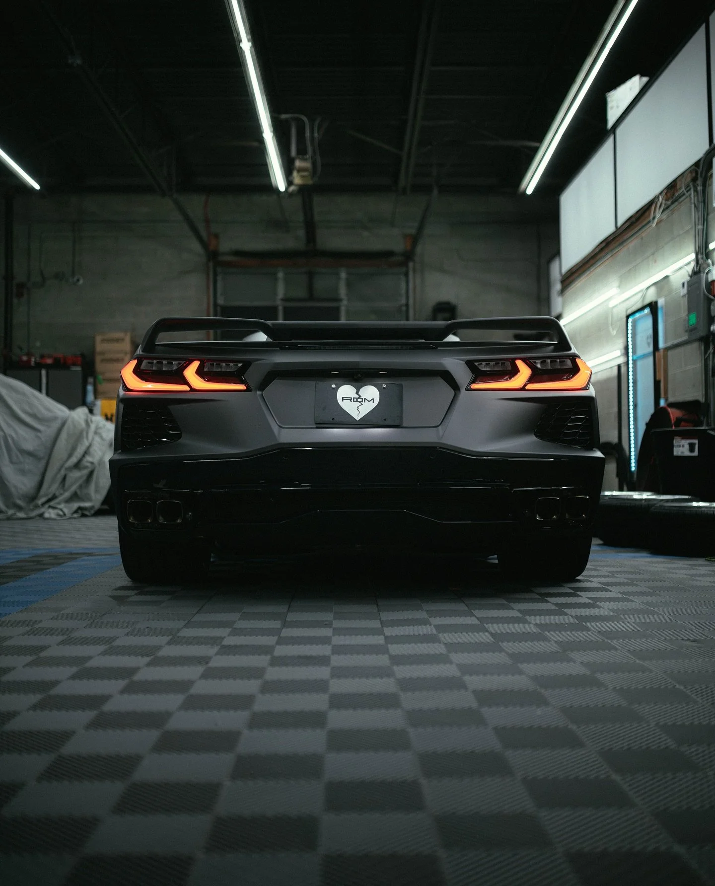 Rear view of a Corvette with a matte gray vinyl wrap with illuminated taillights in a garage, with a checkered floor and various equipment in the background.